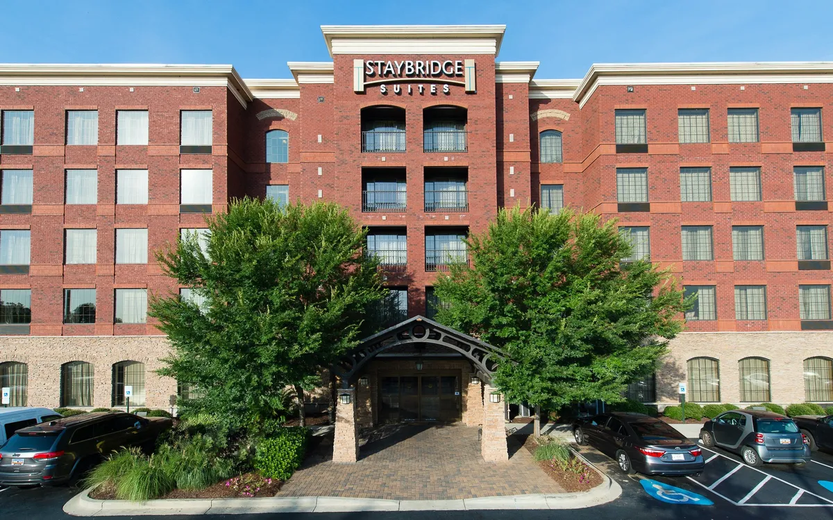 This exterior image showcases the Staybridge Suites' entrance, complete with a prominent sign, a welcoming archway, manicured landscaping, and glimpses of families or guests arriving. The inviting outdoor area hints at the start of an exciting adventure in Columbia.