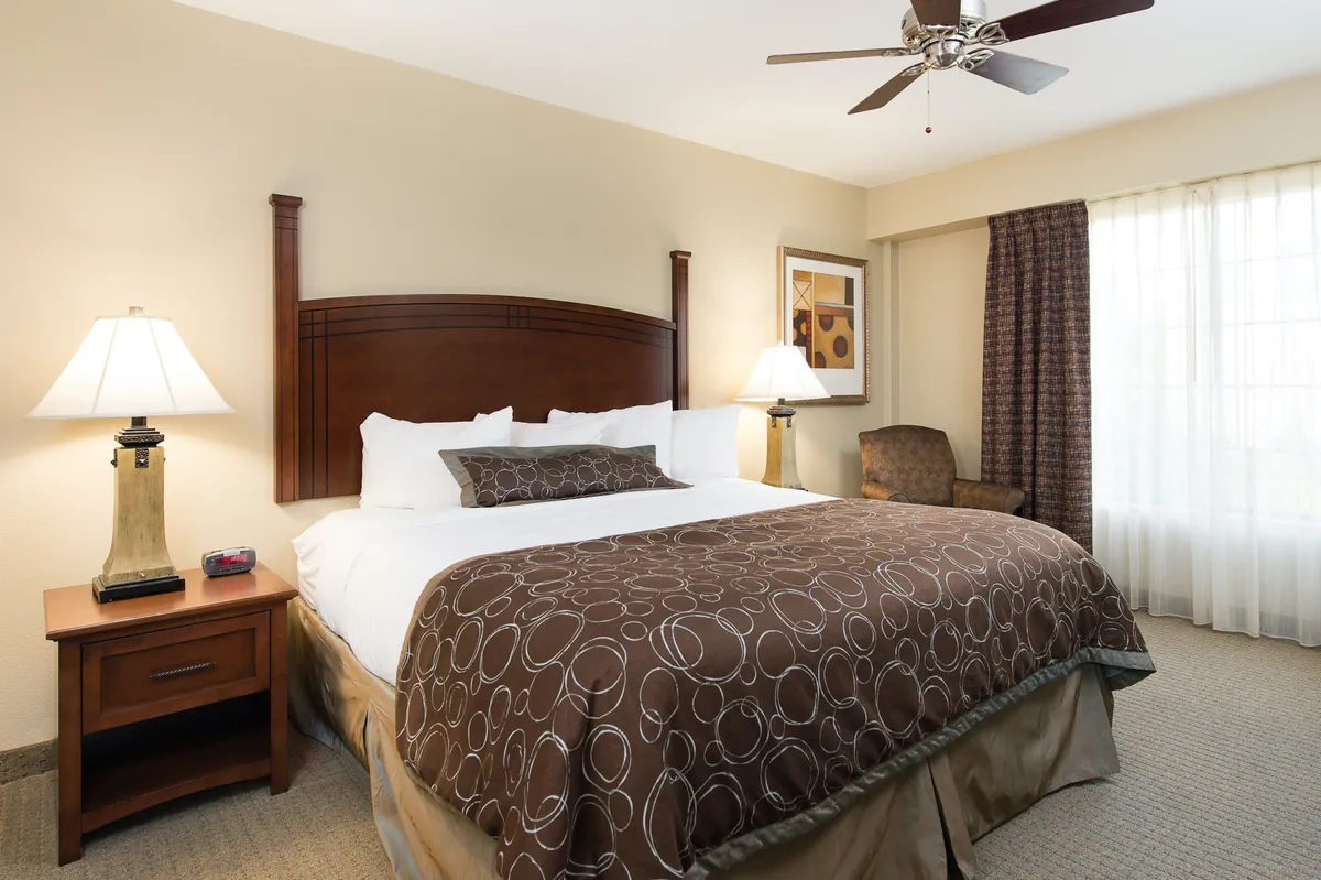 This image features a serene bedroom with a comfortable bed, nightstand, lamps, a cozy chair, and a window allowing in natural light. The soft, neutral decor and plush bedding evoke a sense of relaxation and tranquility, perfect for restful nights after a day of exploration.